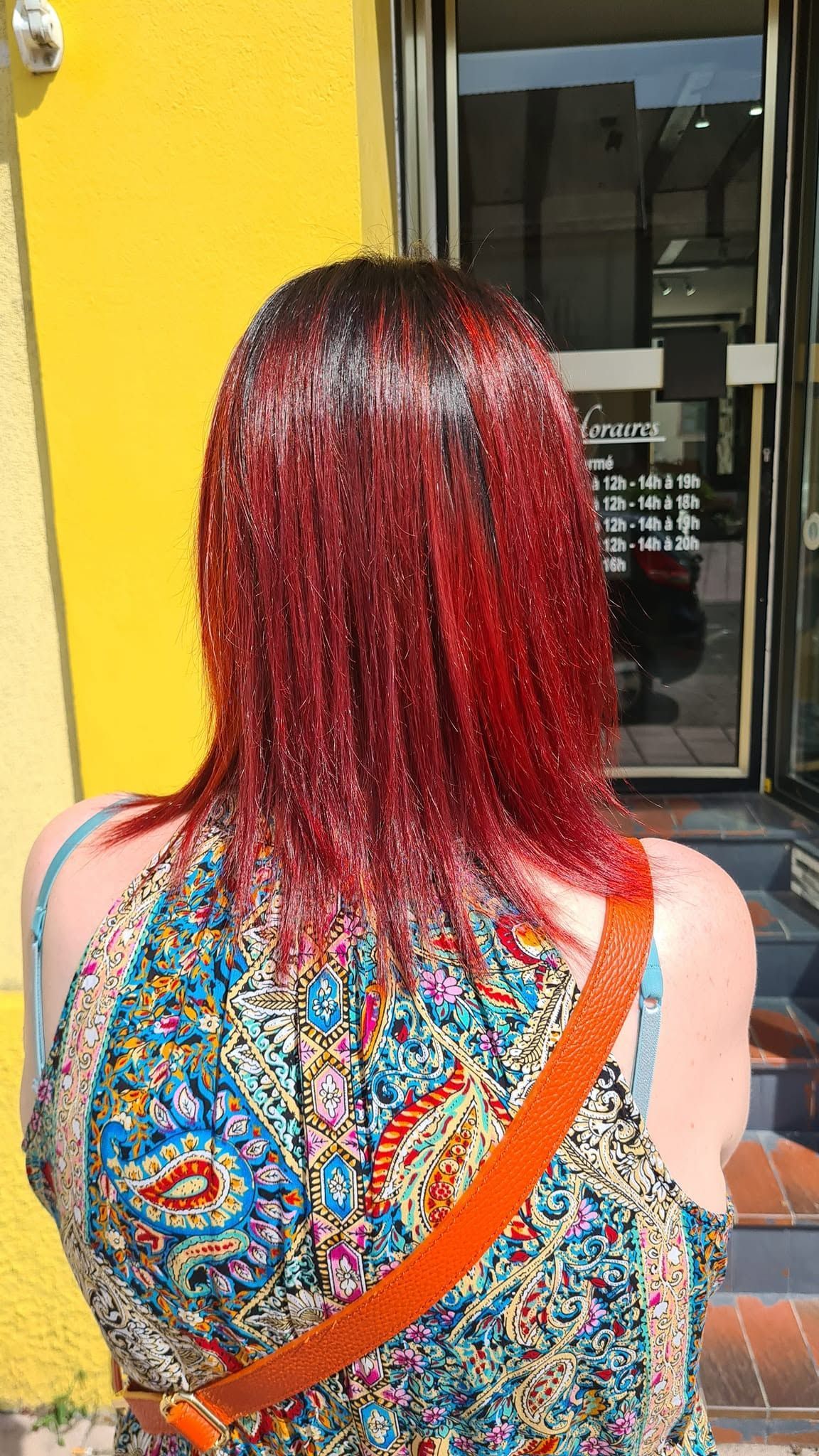 Personne aux cheveux roux vue de dos, portant un haut fleuri coloré à bretelle orange, debout près d'un mur jaune.