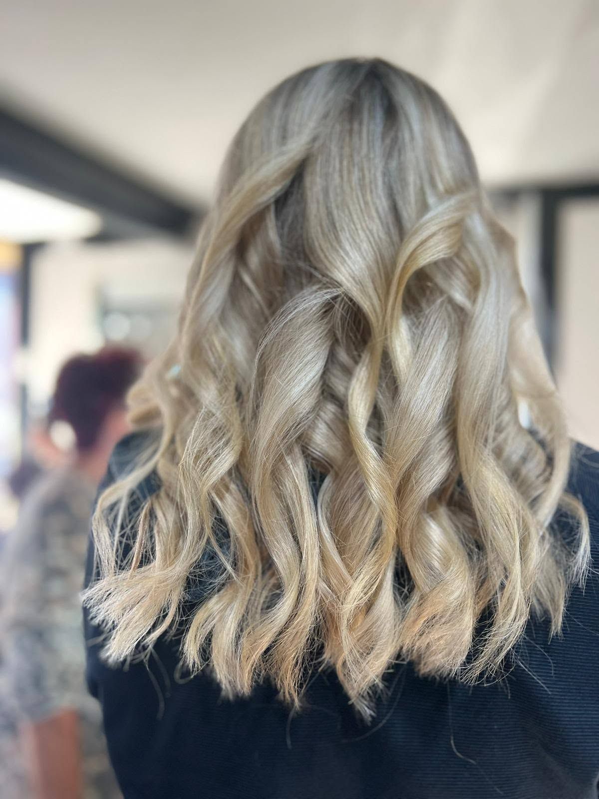 Cheveux blonds ondulés mi-longs avec des racines foncées, vus de dos dans un salon de coiffure.