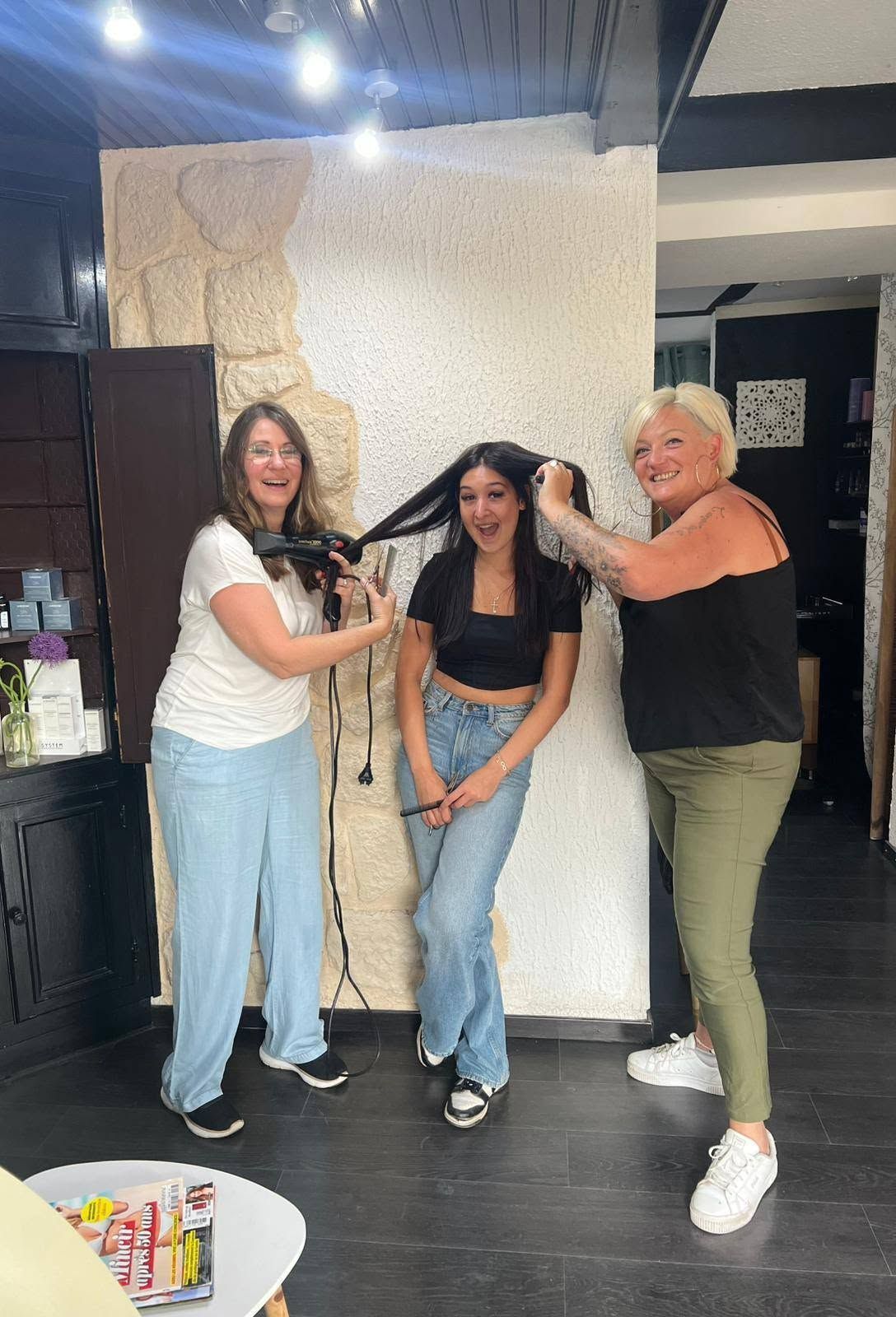 Trois personnes posent ensemble à l'intérieur, souriantes, l'une d'elles ajustant les cheveux de la personne au centre.