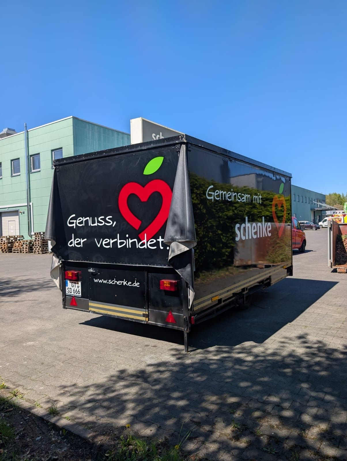 Schwarzer LKW mit rotem Herzlogo und deutschem Text parkt neben Gebäuden an einer sonnigen Straße