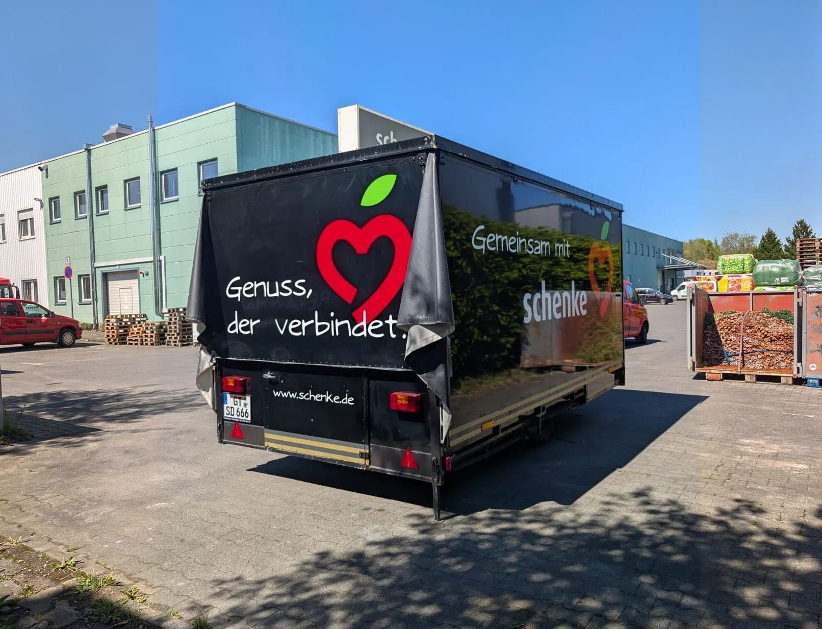 Ein LKW mit dem Herzlogo „Gruß der verbindet“ parkt auf einer sonnigen Straße neben einem Gebäude.