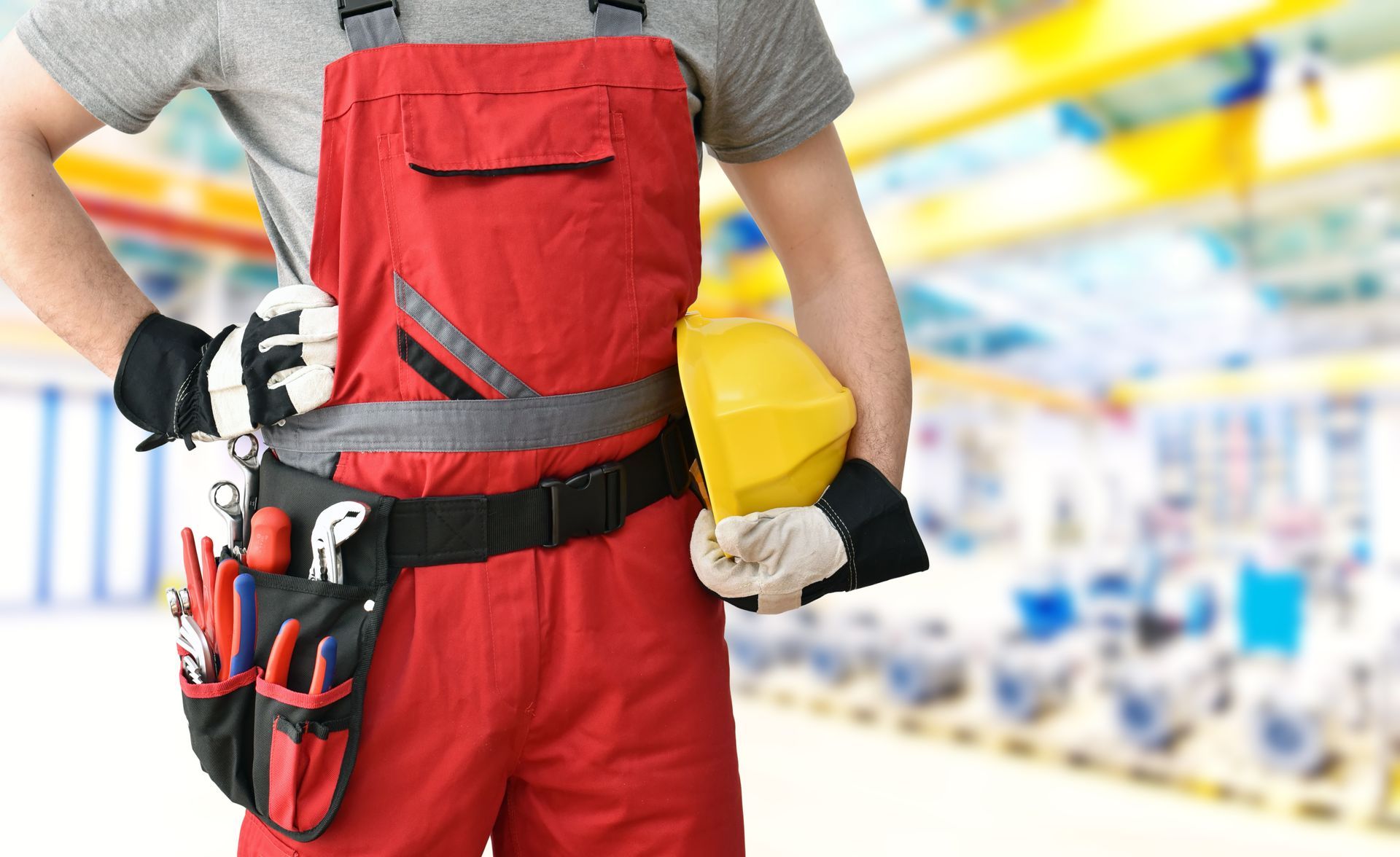 Ein Bauarbeiter in rotem Overall hält einen gelben Schutzhelm auf einer Baustelle und trägt Werkzeug an einem Gürtel.