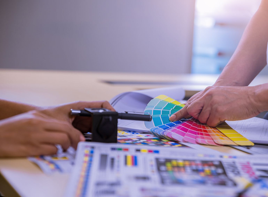 Hände, die einen Farbfächer neben einem Drucker und ausgedruckten Farbkarten auf einem Schreibtisch auswählen