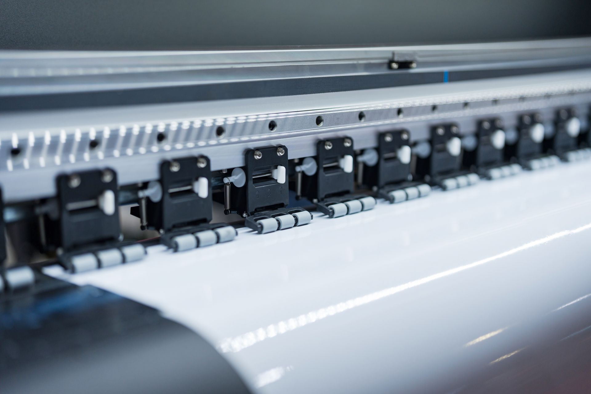 Nahaufnahme einer Textilmaschine mit schwarzen Klammern und grauen Bauteilen, die weißen Stoff zuführen.
