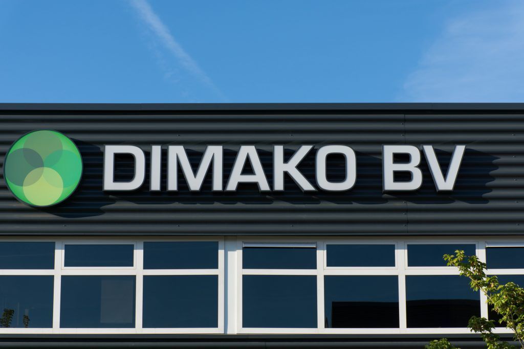 DIMAKO BV-Gebäudeschild mit grünem, kreisförmigem Logo an einer modernen Fassade unter blauem Himmel