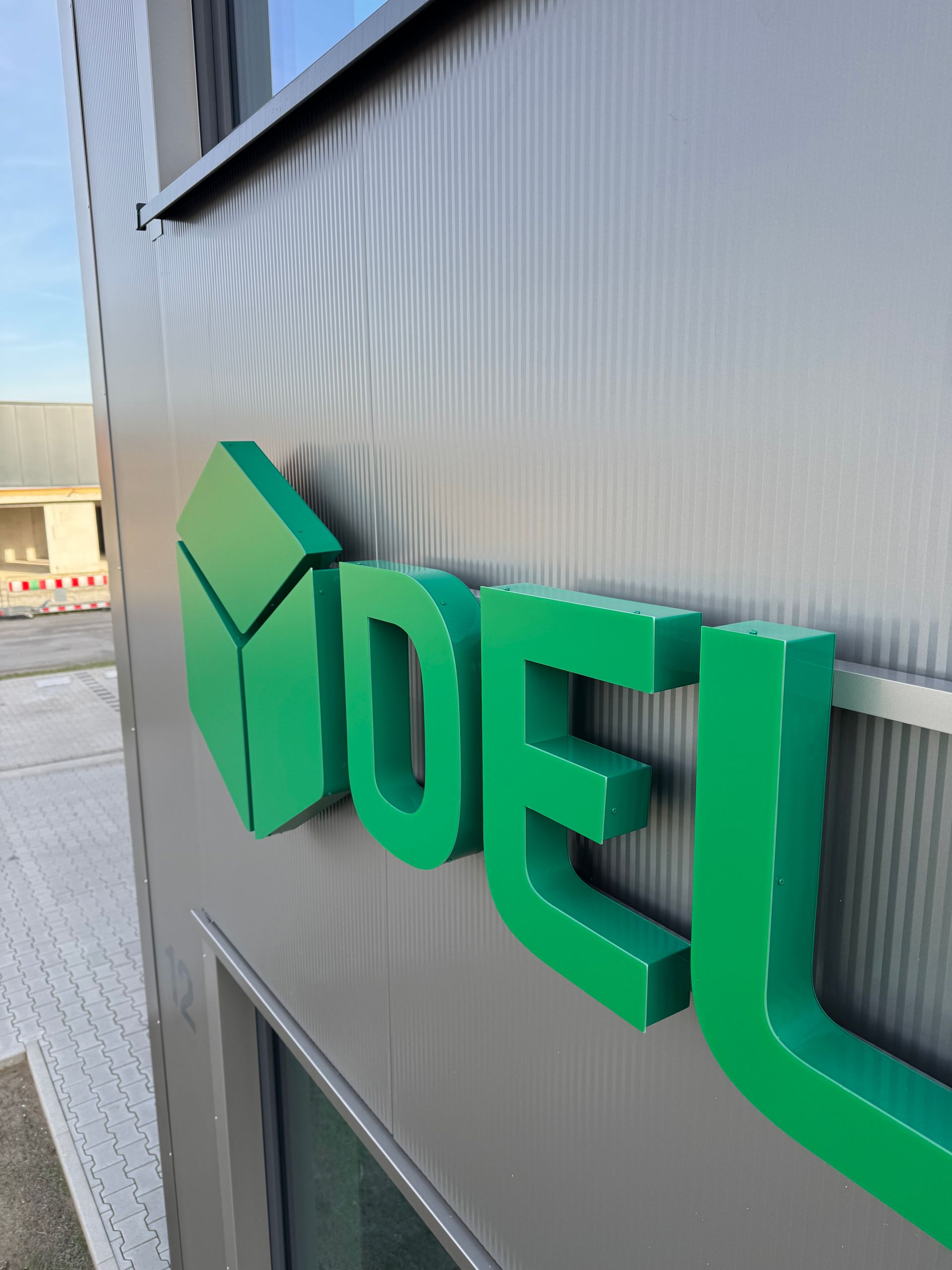 Grünes 3D-Firmenlogo auf der Außenfassade eines Wellblechgebäudes, aus einem bestimmten Winkel betrachtet
