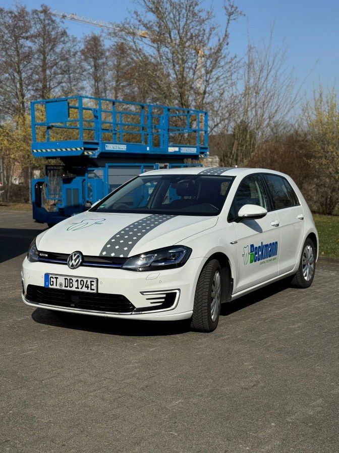 Ein weißer VW-Pkw parkt neben einer blauen Hubarbeitsbühne an einer Straße; im Hintergrund sind Bäume zu sehen.