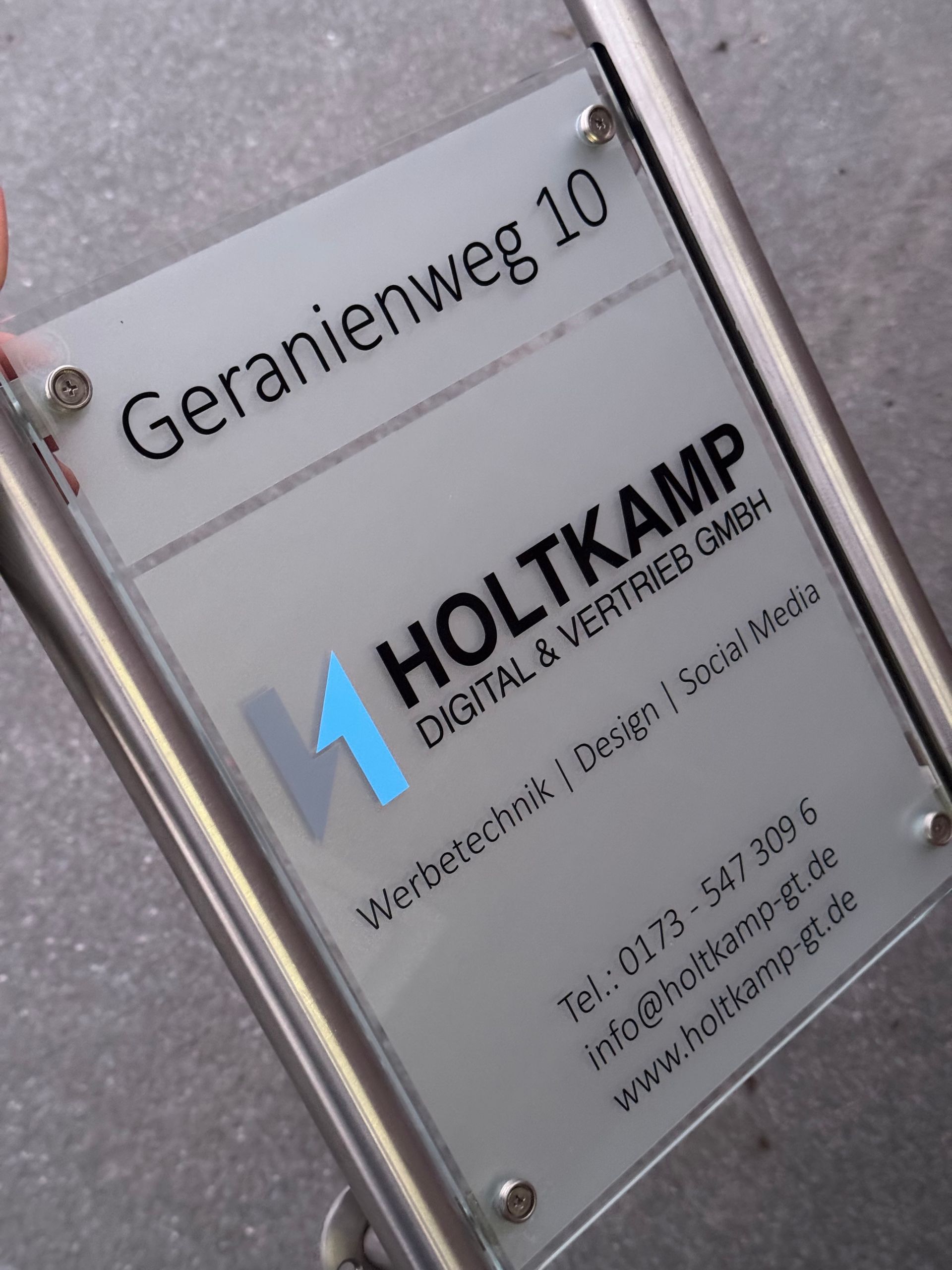 Ein Straßenschild mit der Aufschrift „Geranienweg 10“ neben einer digitalen Servicetafel von HOLTKAMP.