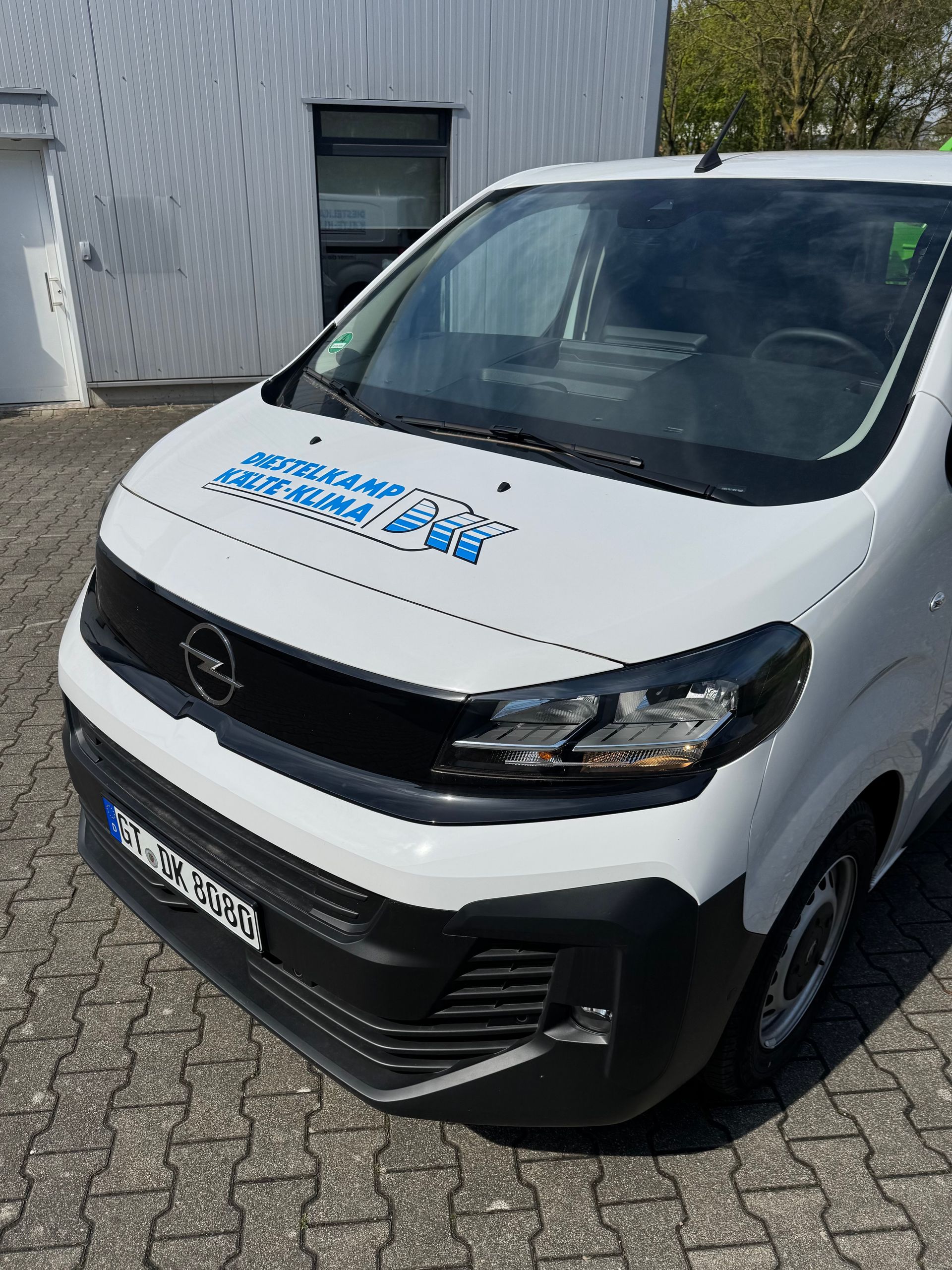 Weißer Lieferwagen parkt vor einem Gebäude, Vorderansicht mit blauem Logo auf der Motorhaube.