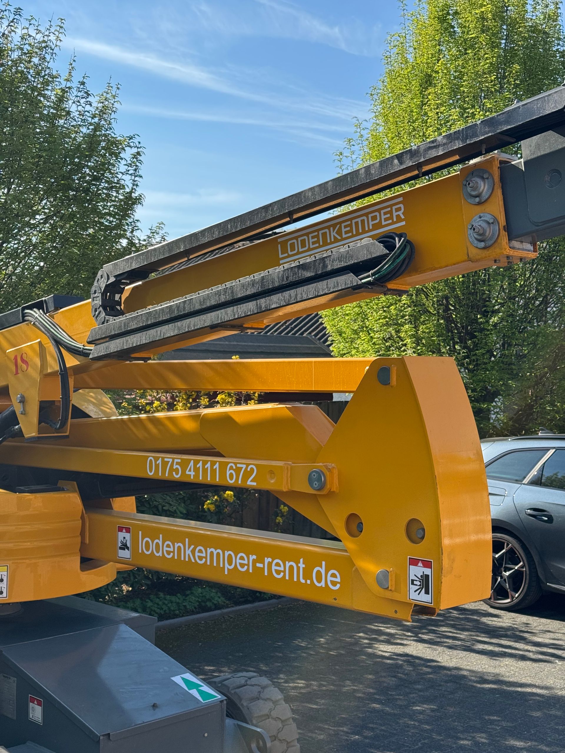 Gelber Kranarm mit Sägeaufsatz auf einer Baustelle im Freien, im Hintergrund Bäume.