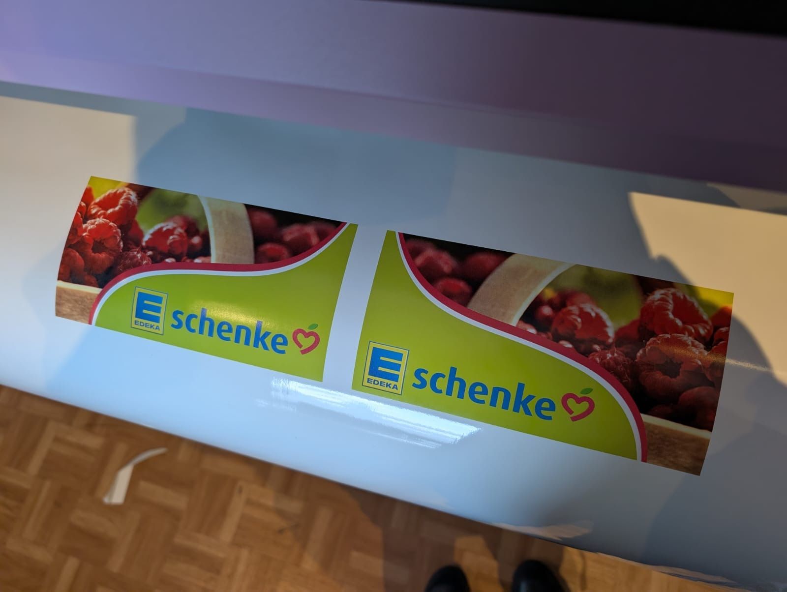 Zwei farbenfrohe Schenke-Poster mit Fruchtmotiven hängen an einer Wand über einem Parkettboden.