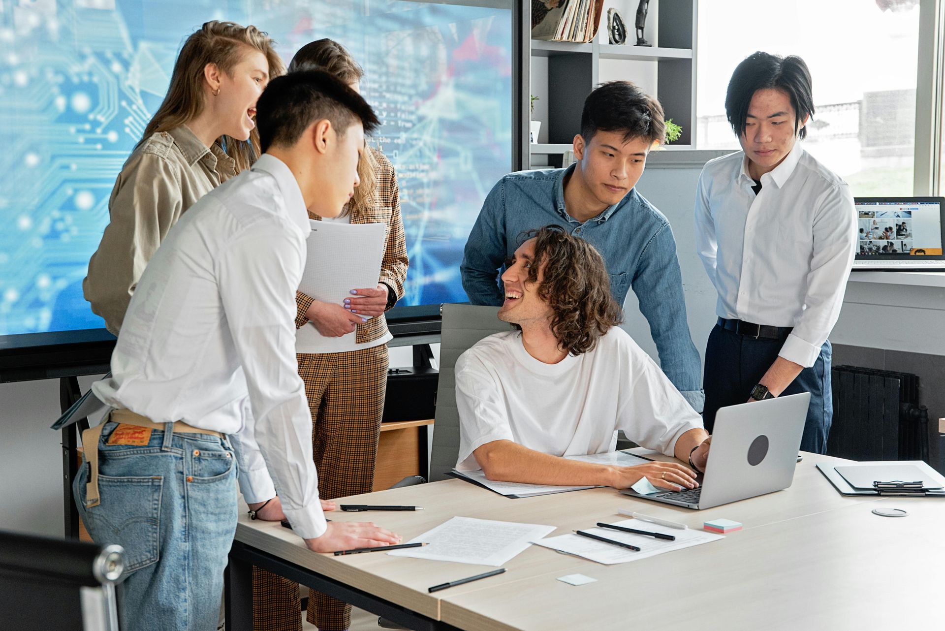 Ein Team arbeitet in einem hellen Büro an einem Schreibtisch zusammen; im Hintergrund sind ein Laptop, Papiere und ein Whiteboard zu sehen.