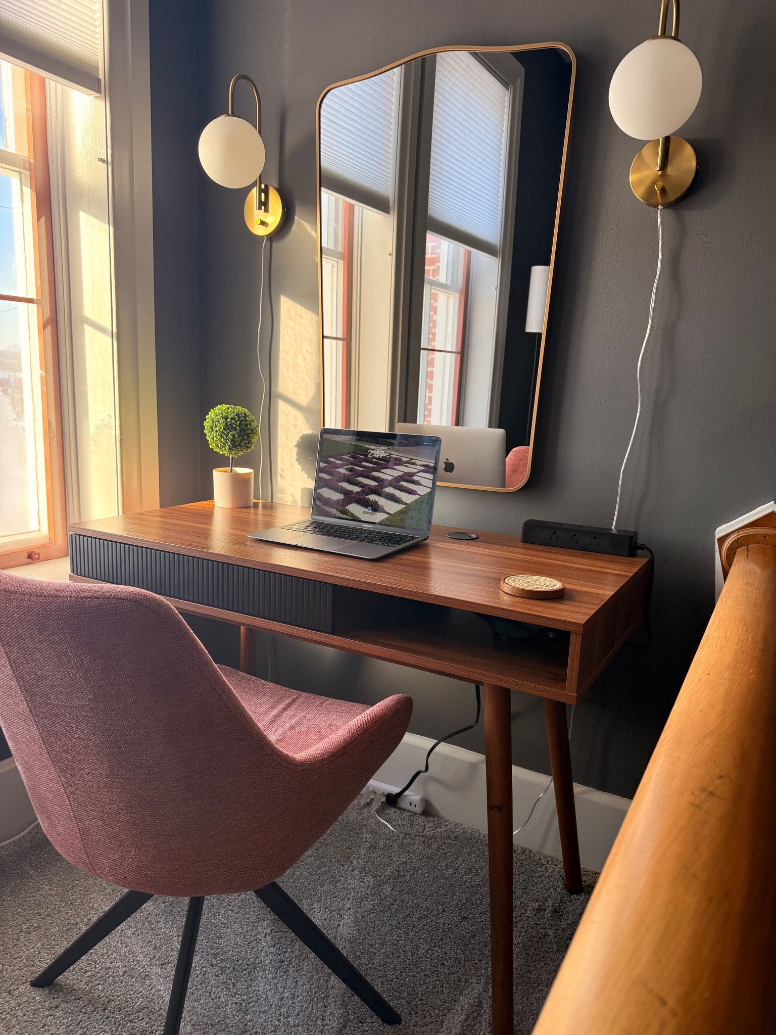Cozy vanity desk by a window, with pink chair, mirror, wall sconces, and small plant.