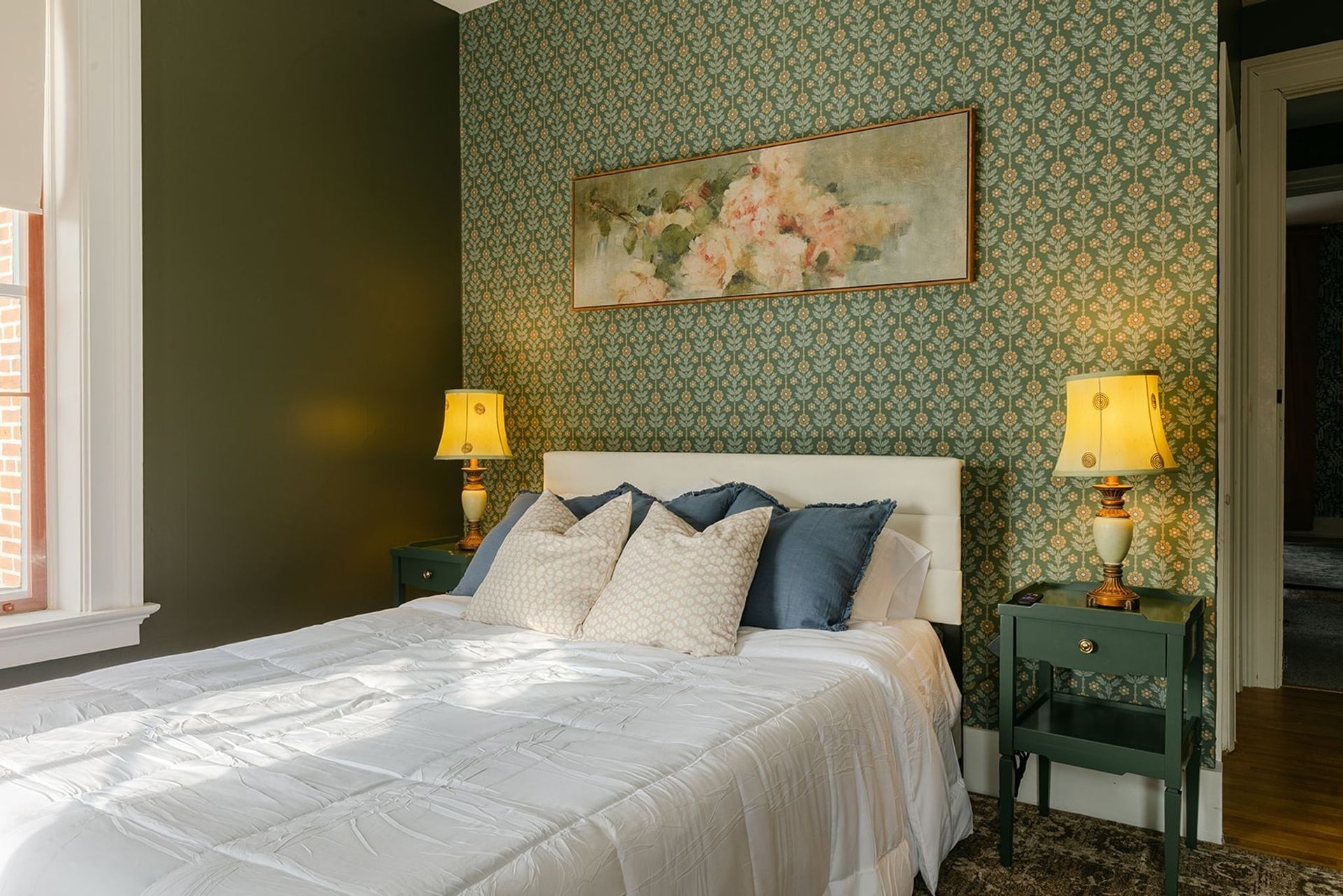 Sunlit bedroom with green patterned wallpaper, white bed, floral art, and yellow lamps on both sides