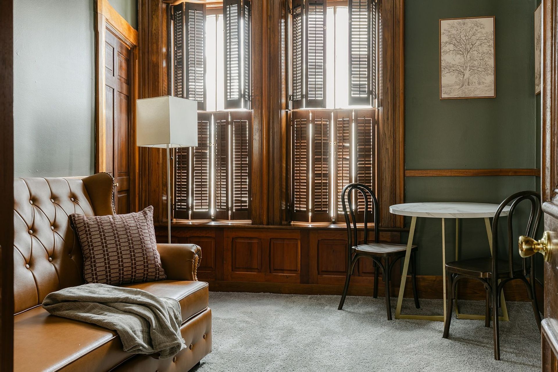 Vintage sitting room with tufted chair, window shutters, and small round table by a cane chair