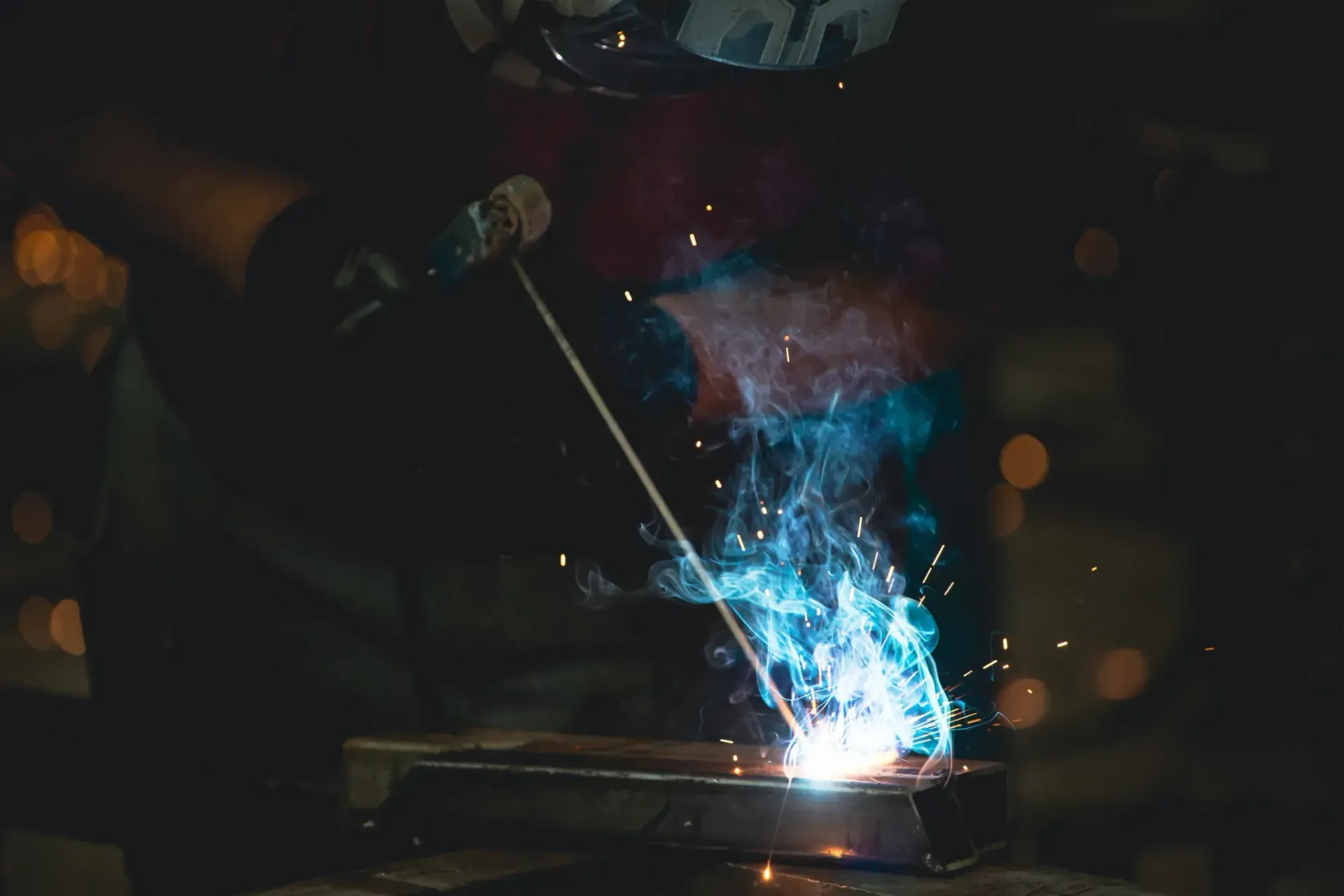 Soldador en una habitación oscura utilizando un soplete de soldadura, chispas azules brillantes y humo.