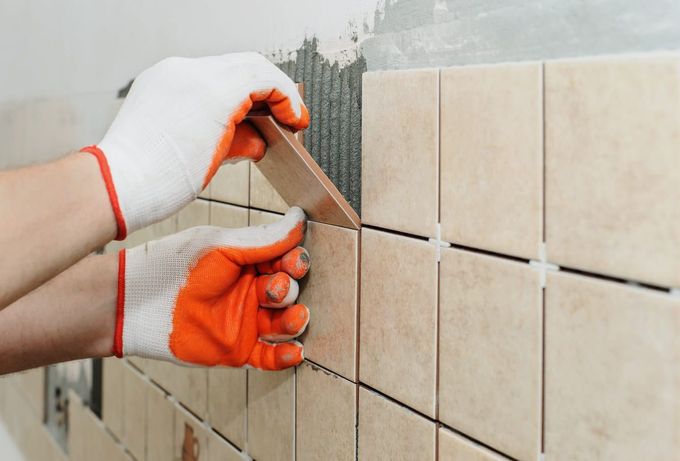 Persona con guantes instalando baldosas de color beige en una pared, utilizando espaciadores para baldosas.