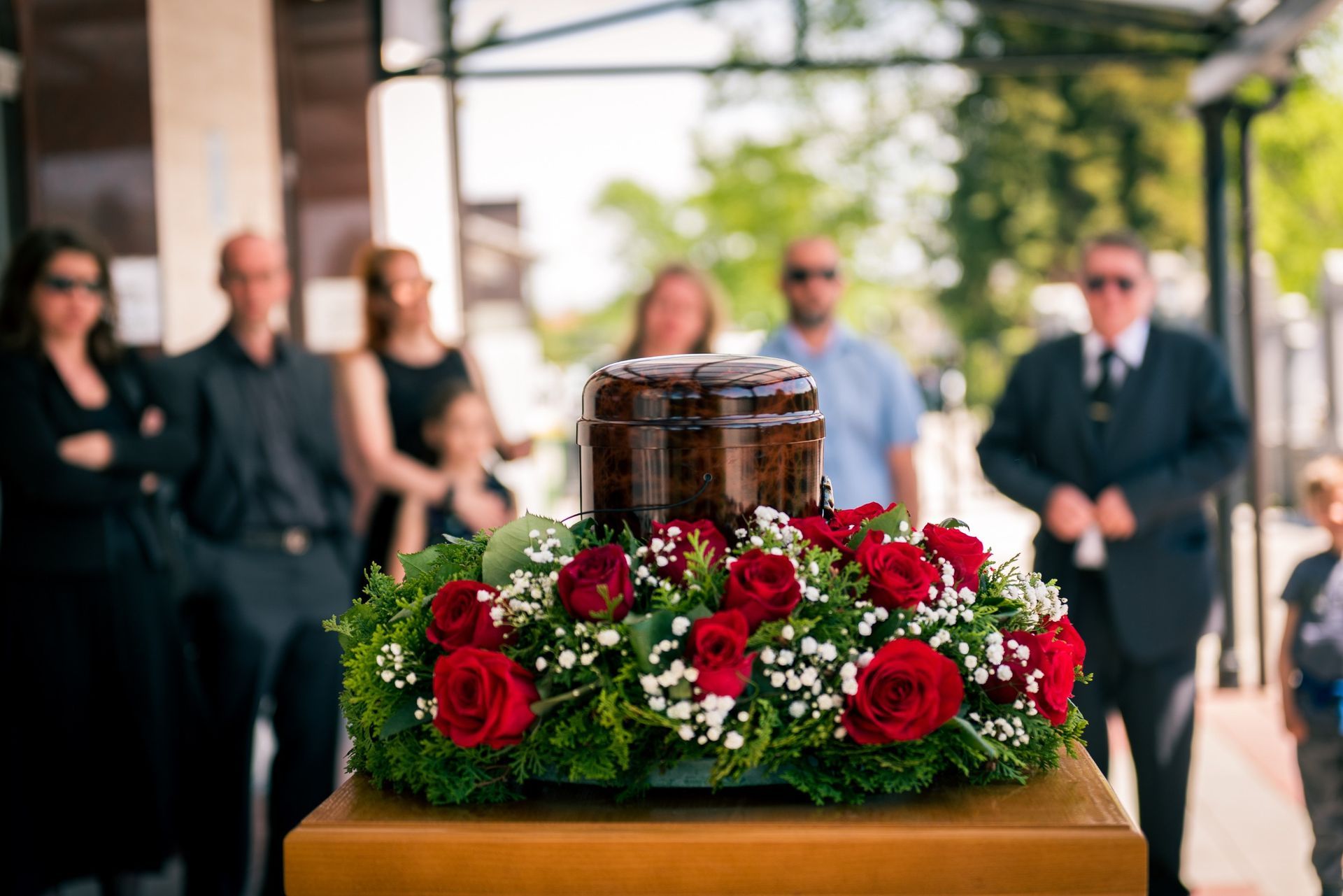 Urna sobre una mesa de madera con una corona de rosas rojas, rodeada por un grupo de personas vestidas de negro en un servicio conmemorativo al aire libre.