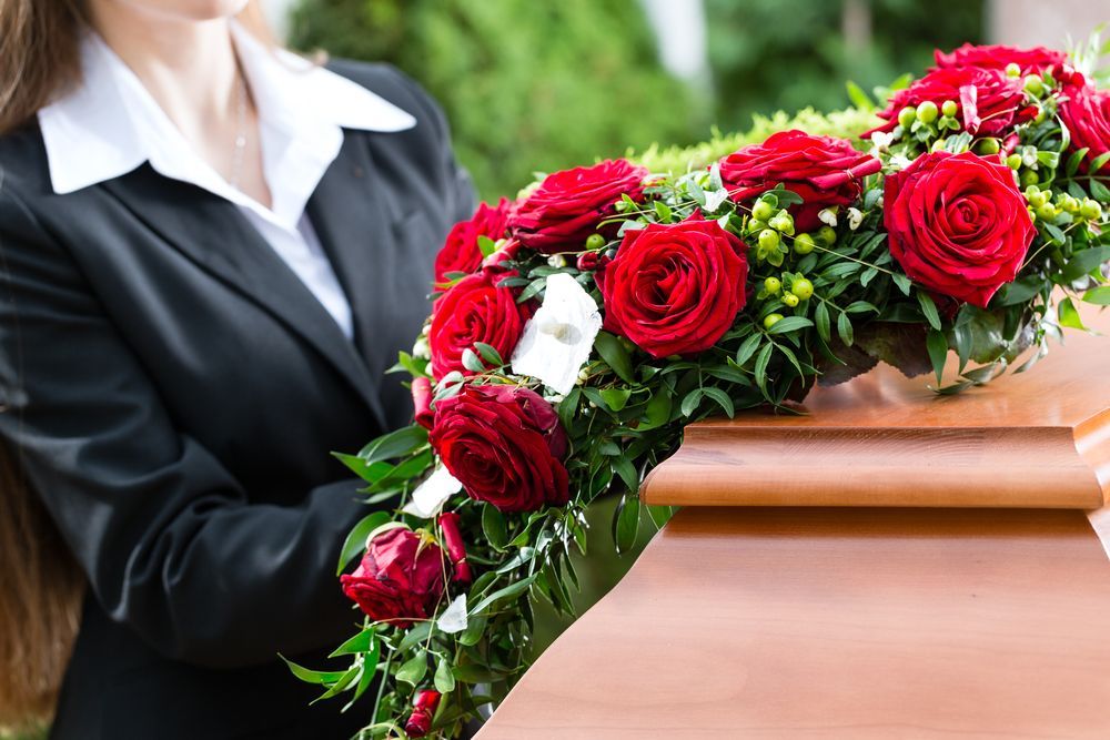 Mujer con traje negro sosteniendo un arreglo floral de rosas rojas sobre un ataúd de madera en un funeral.