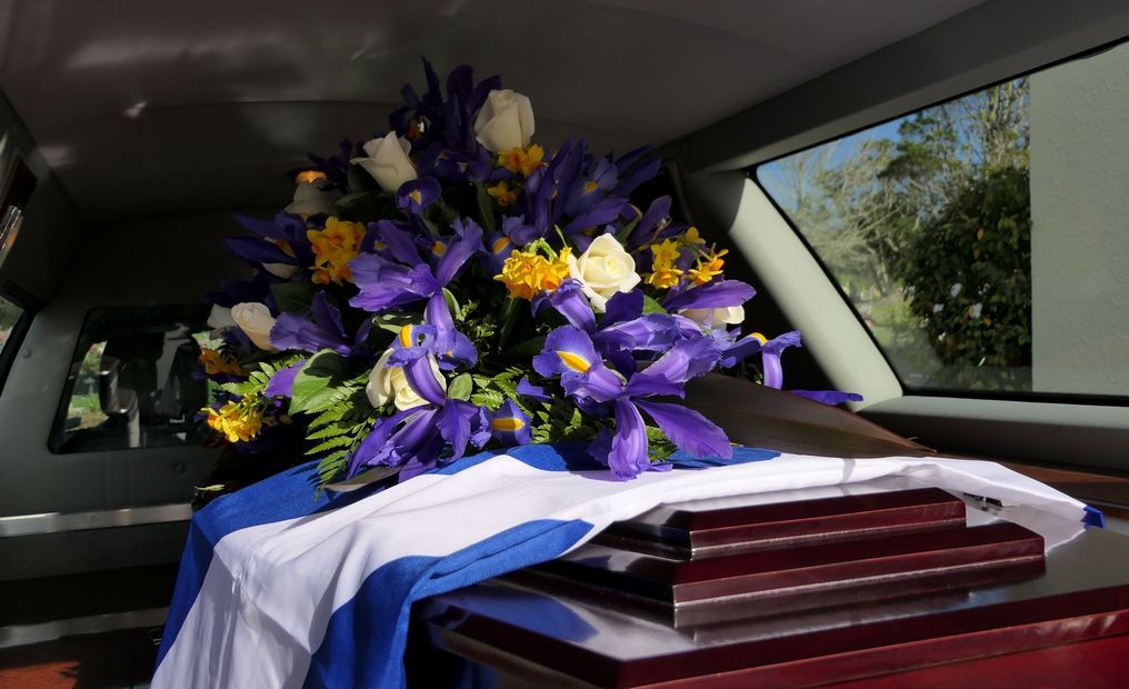 Un ataúd cubierto con una bandera azul y blanca dentro de un coche fúnebre, coronado con un arreglo floral de flores violetas, blancas y amarillas.