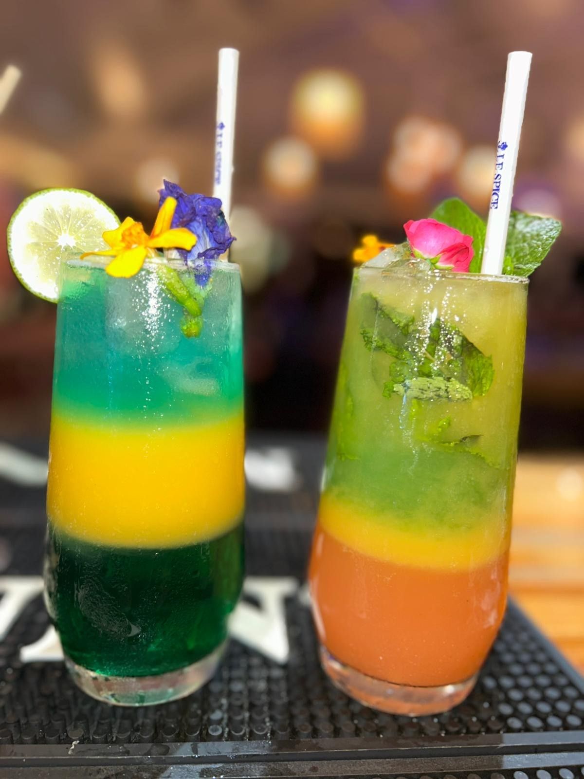 Deux cocktails superposés garnis de fleurs, de citron vert et de menthe ; l'un bleu/jaune/vert, l'autre vert/jaune/orange.