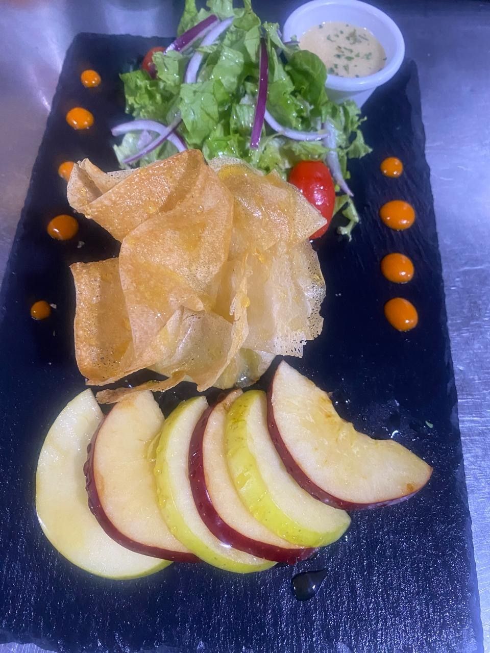 Salade avec tranches de pommes, frites, légumes verts, oignons rouges, tomates, vinaigrette, sur une assiette rectangulaire noire.