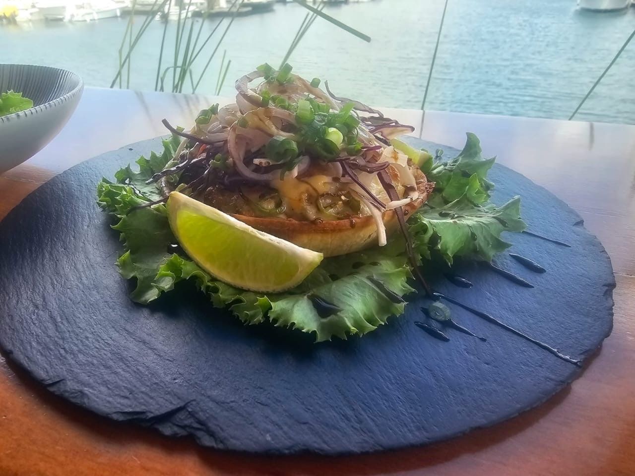 Plat sur une assiette sombre, garni de légumes et d'un quartier de citron vert. Marina floue en arrière-plan.