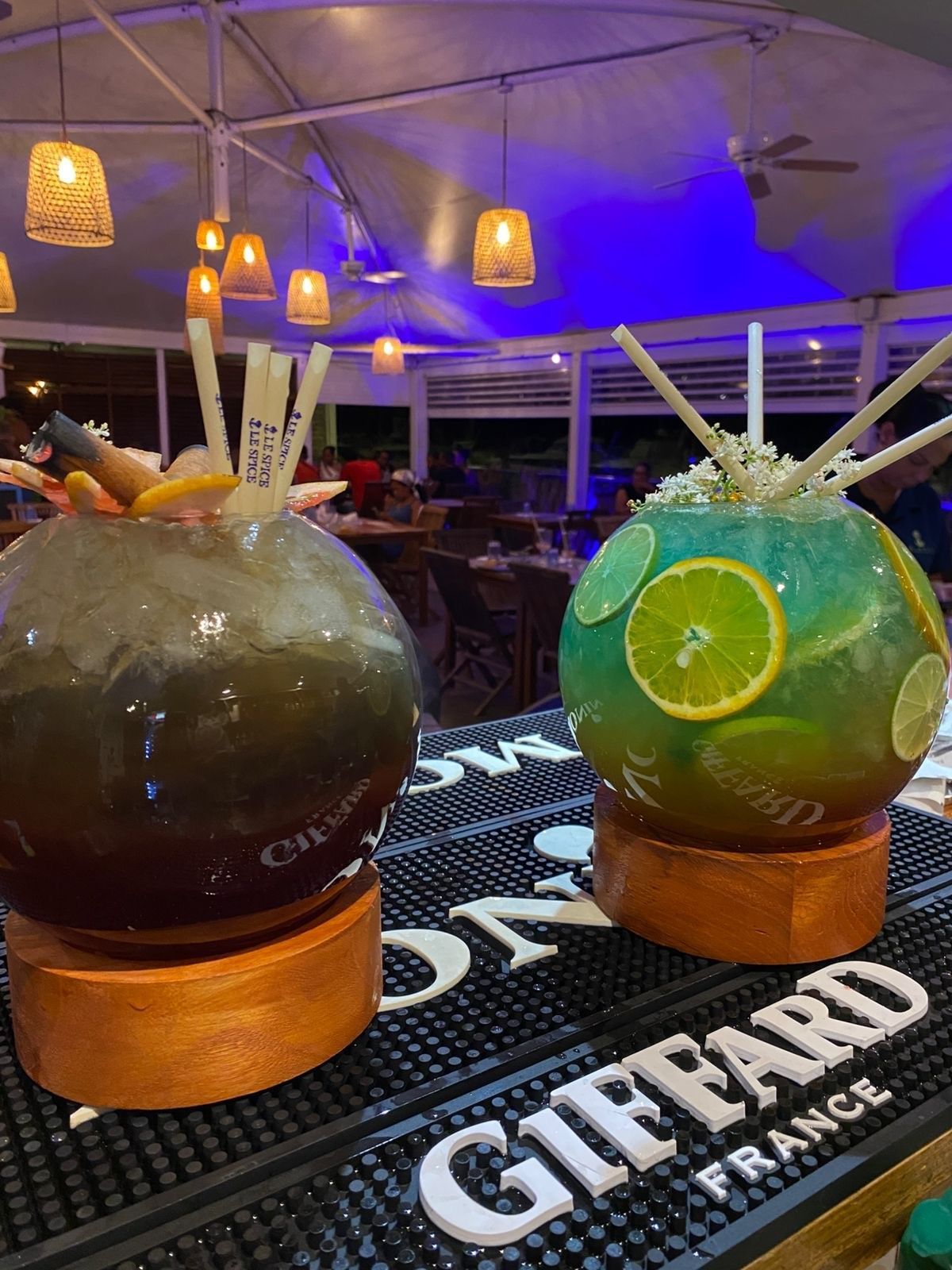 Deux grands cocktails dans des sphères décoratives sur un bar, l'un foncé, l'autre vert, avec des pailles et des tranches de fruits.