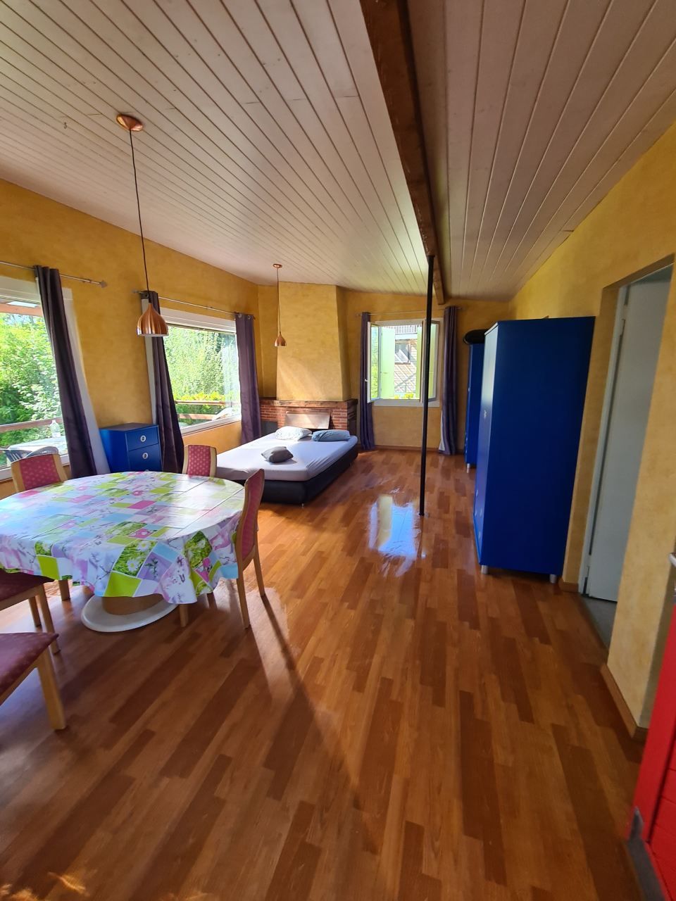 Une pièce lumineuse aux murs jaunes, au parquet, avec une table à manger, un lit et une grande armoire.