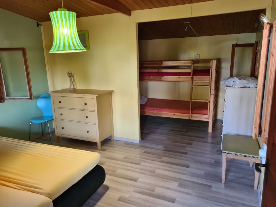 Une chambre avec un lit double jaune, une commode en bois, une lampe suspendue verte.