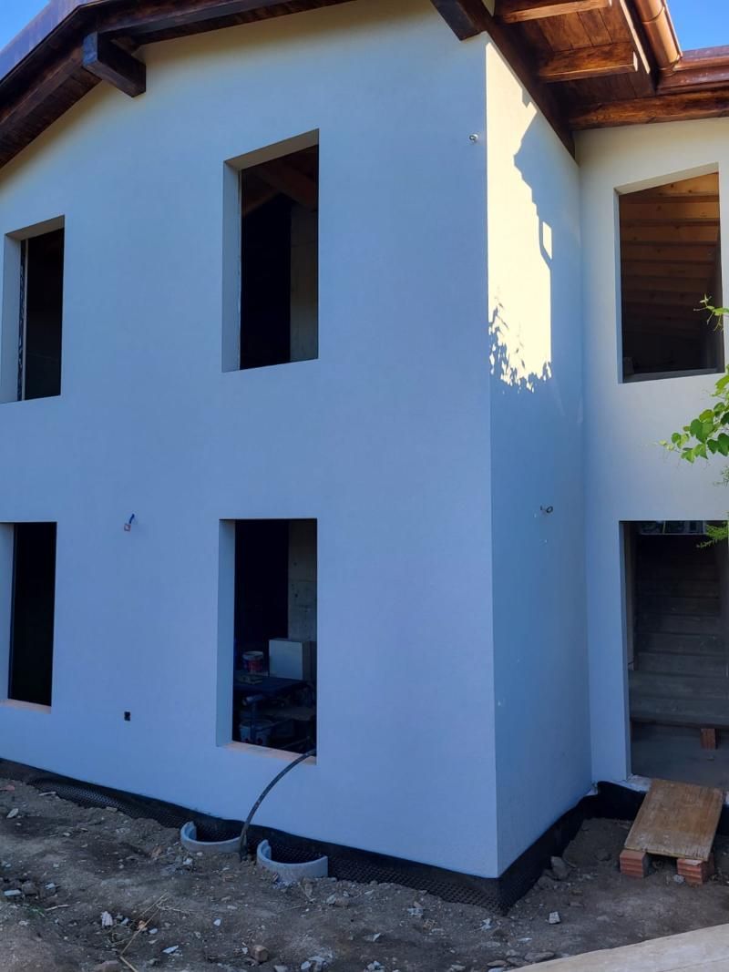 Esterno di una casa a due piani in costruzione; pareti di colore chiaro, aperture per finestre e travi del tetto marroni.