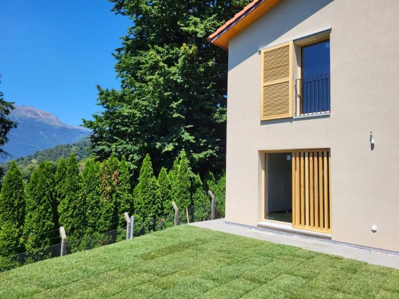 Casa beige con persiane e porta gialle, prato verde, vista sulle montagne, cielo azzurro.
