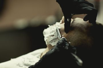 Una mano enguantada afeitando el rostro de una persona con una navaja de afeitar, primer plano en un entorno con poca luz.