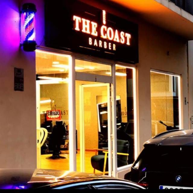Exterior nocturno de The Coast Barbers, con entrada iluminada con luz cálida y un coche aparcado delante.