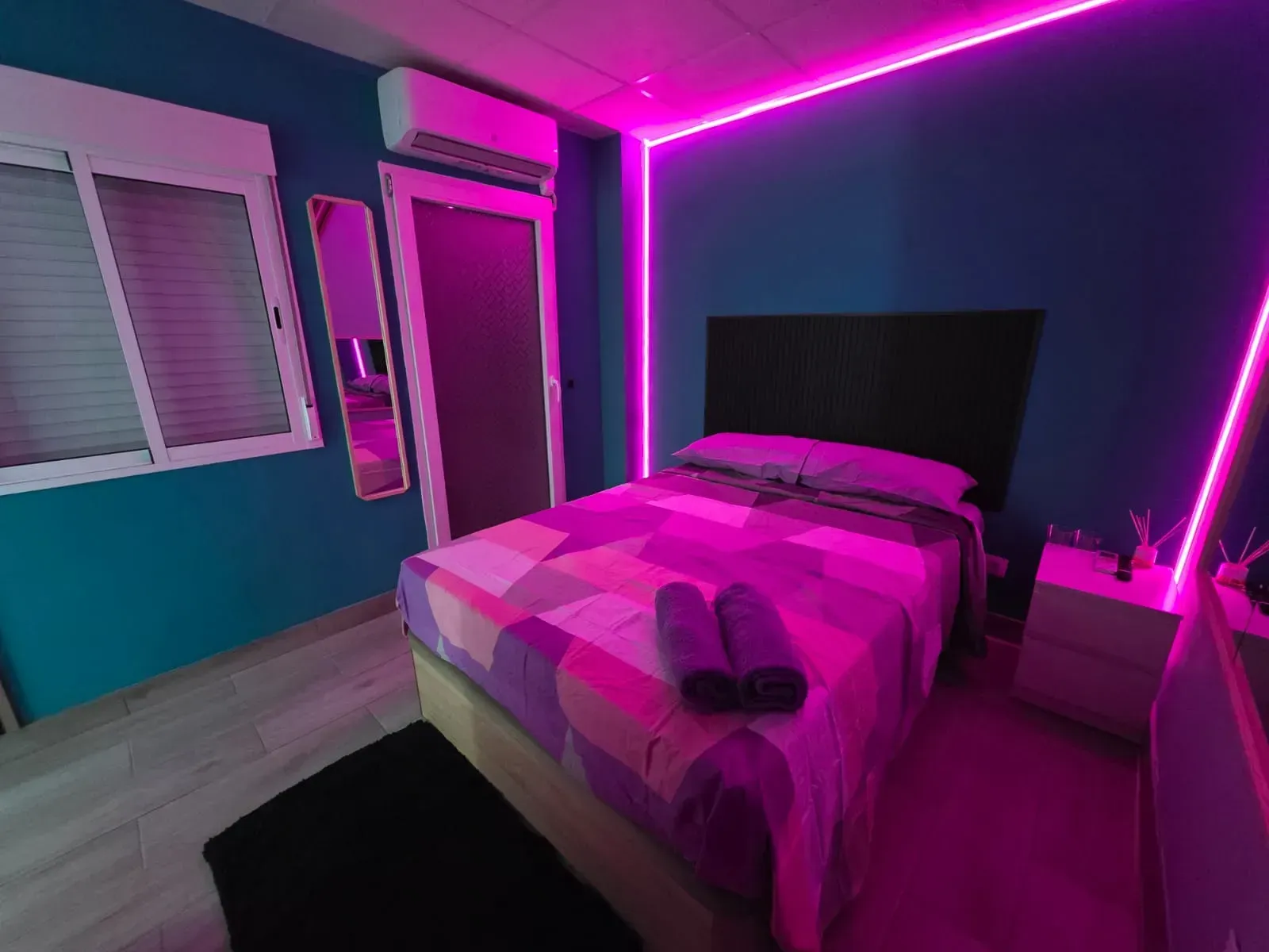 Un dormitorio con cama, mesita de noche, espejo y luces violetas.
