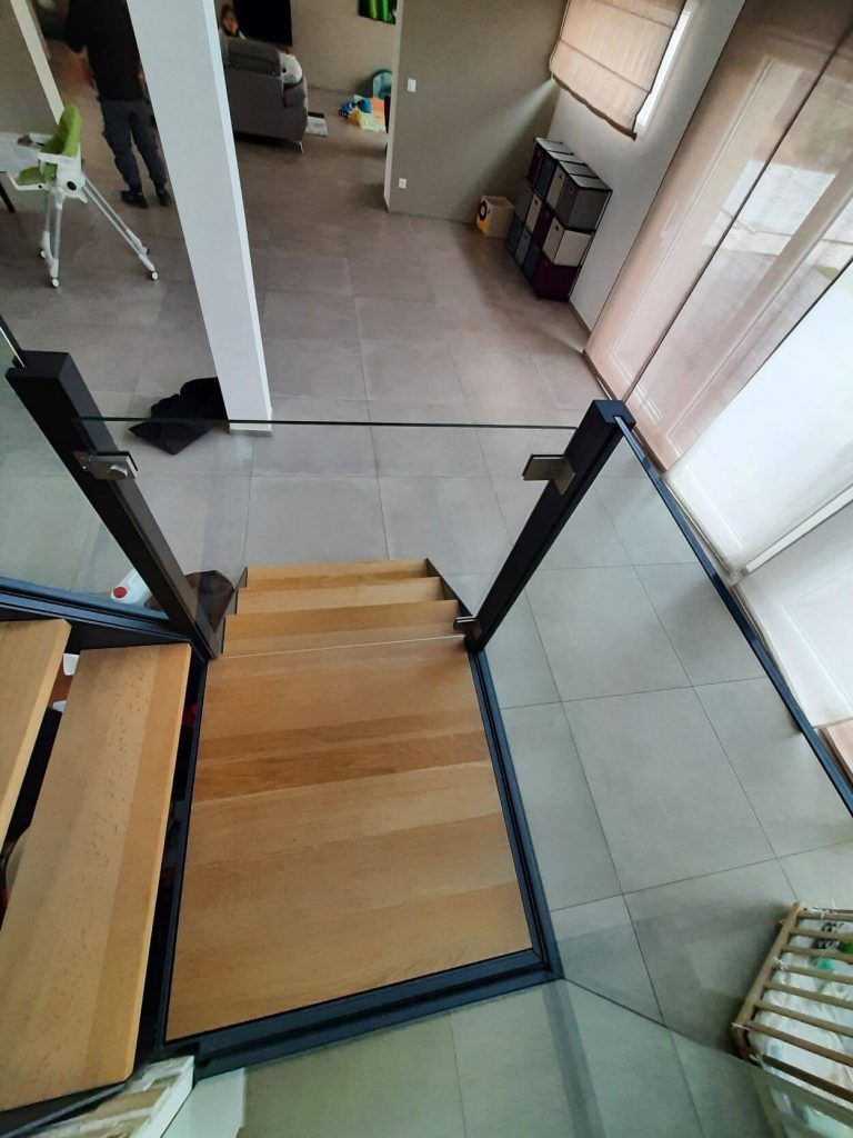 Vue en plongée sur un escalier en bois avec des rampes noires et des panneaux de verre donnant sur un espace de vie moderne carrelé.