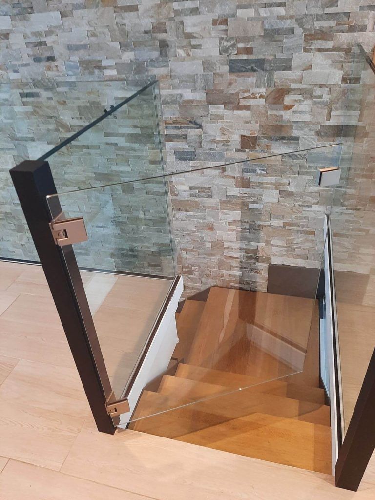Vue en plongée d'un escalier en bois descendant, entouré de balustrades noires modernes avec des panneaux de verre, adossé à un mur en pierre.