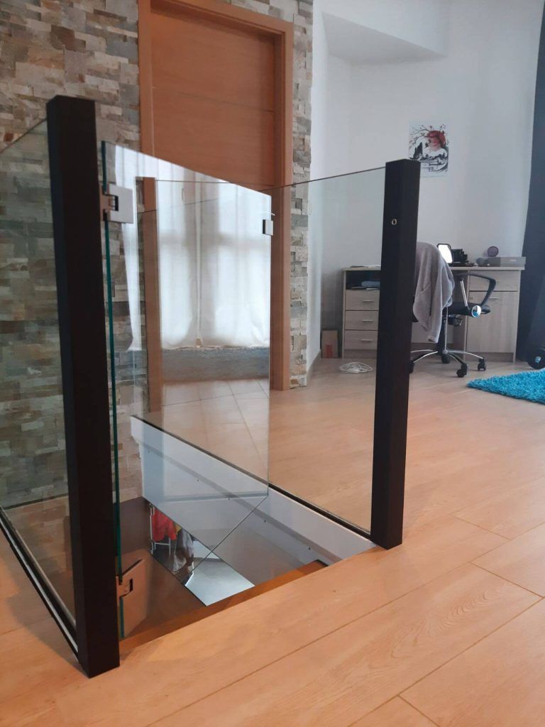 Une balustrade en verre avec des poteaux en bois noir borde un escalier dans une pièce au parquet clair et au mur orné de pierres.