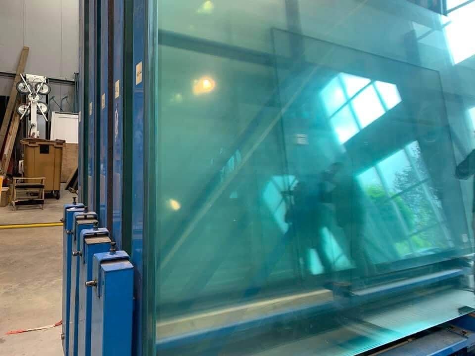 De grandes plaques de verre teintées turquoise sont stockées verticalement sur un support métallique dans un espace de travail industriel.