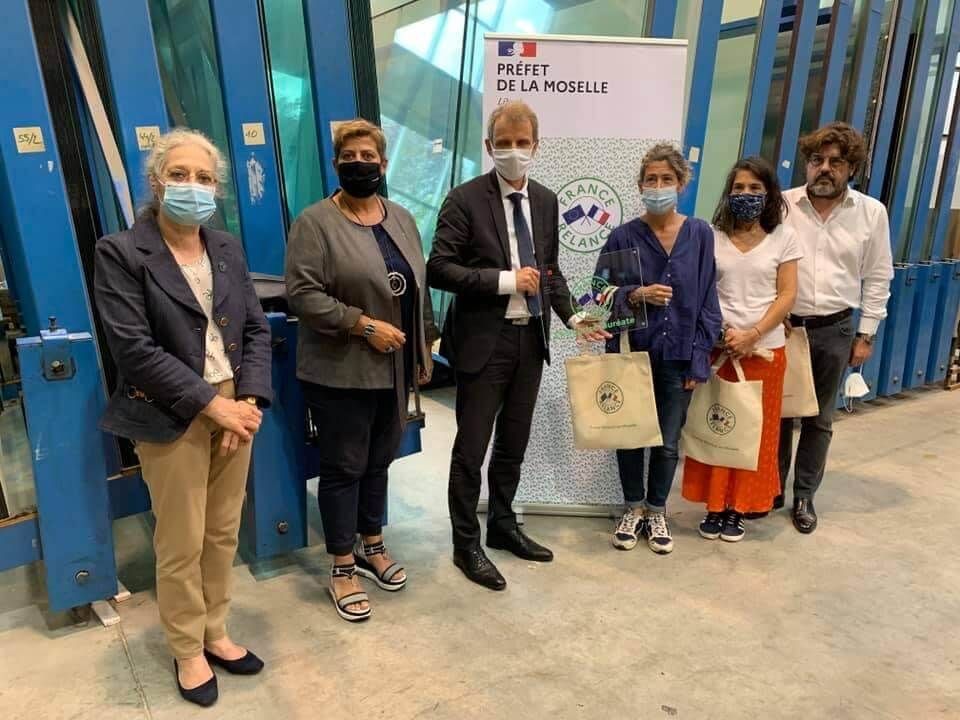 Six personnes se tiennent en rang lors d'une présentation devant un panneau du préfet de Moselle, tenant des sacs fourre-tout et une plaque.