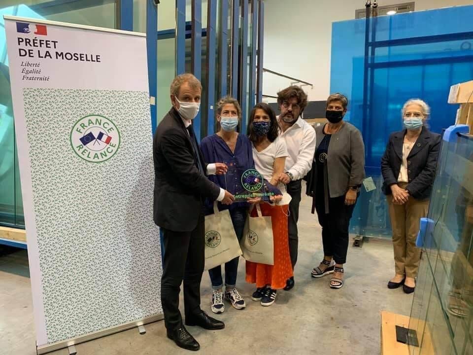 Dans un atelier industriel, à côté d'un panneau d'affichage de la marque, un groupe de personnes portant des masques brandit un prix France Relance.