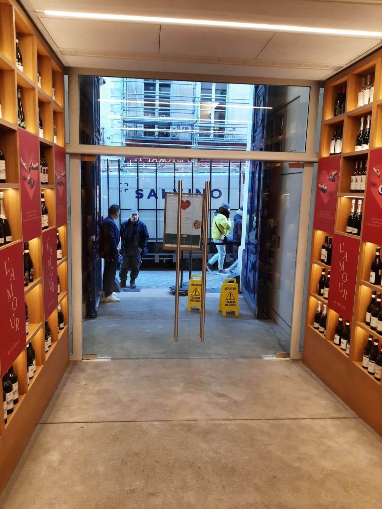 Vue depuis un magasin présentant des bouteilles de vin sur des étagères en bois, donnant sur une rue animée avec des passants et un camion de chantier.