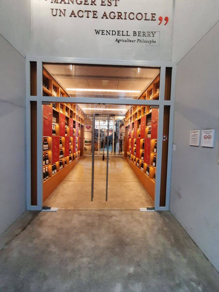 L'entrée d'un magasin, illuminée de façon lumineuse, présente des étagères à vin le long des deux murs et une citation de Wendell Berry imprimée au-dessus de la porte.