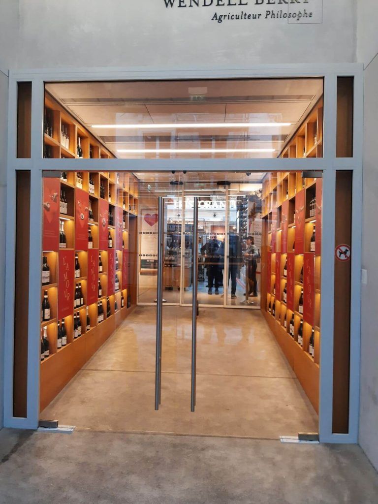Une entrée moderne en verre donne sur un couloir bordé d'étagères en bois remplies de bouteilles de vin, sur fond de panneaux rouges.