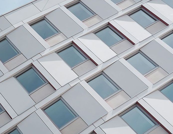 Vue en contre-plongée de la façade d'un bâtiment moderne présentant un motif répétitif de panneaux gris clair et de fenêtres rectangulaires.