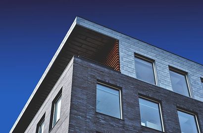 Vue en contre-plongée d'un bâtiment moderne en briques grises, bardage métallique et balcon lambrissé sous un ciel dégagé.