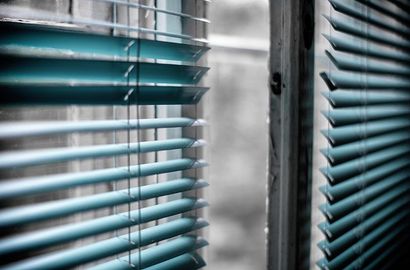 Gros plan sur des stores horizontaux turquoise partiellement baissés, révélant une vue floue à travers le cadre de la fenêtre.