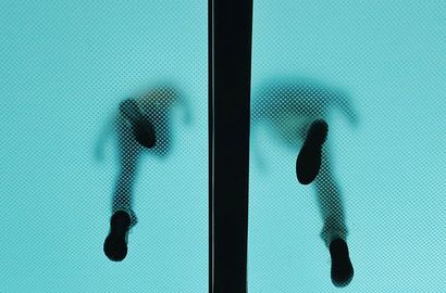 Vue en contre-plongée de deux personnes marchant sur un sol en verre translucide aux tons turquoise, avec une poutre structurelle sombre au centre.