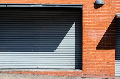 Une porte métallique grise enroulable, fermée, encastrée dans un mur de briques orange, sur laquelle se projette une ombre triangulaire.