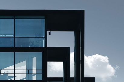 Vue minimaliste d'un bâtiment moderne aux fenêtres en verre bleu et aux cadres architecturaux noirs se détachant sur un ciel nuageux.
